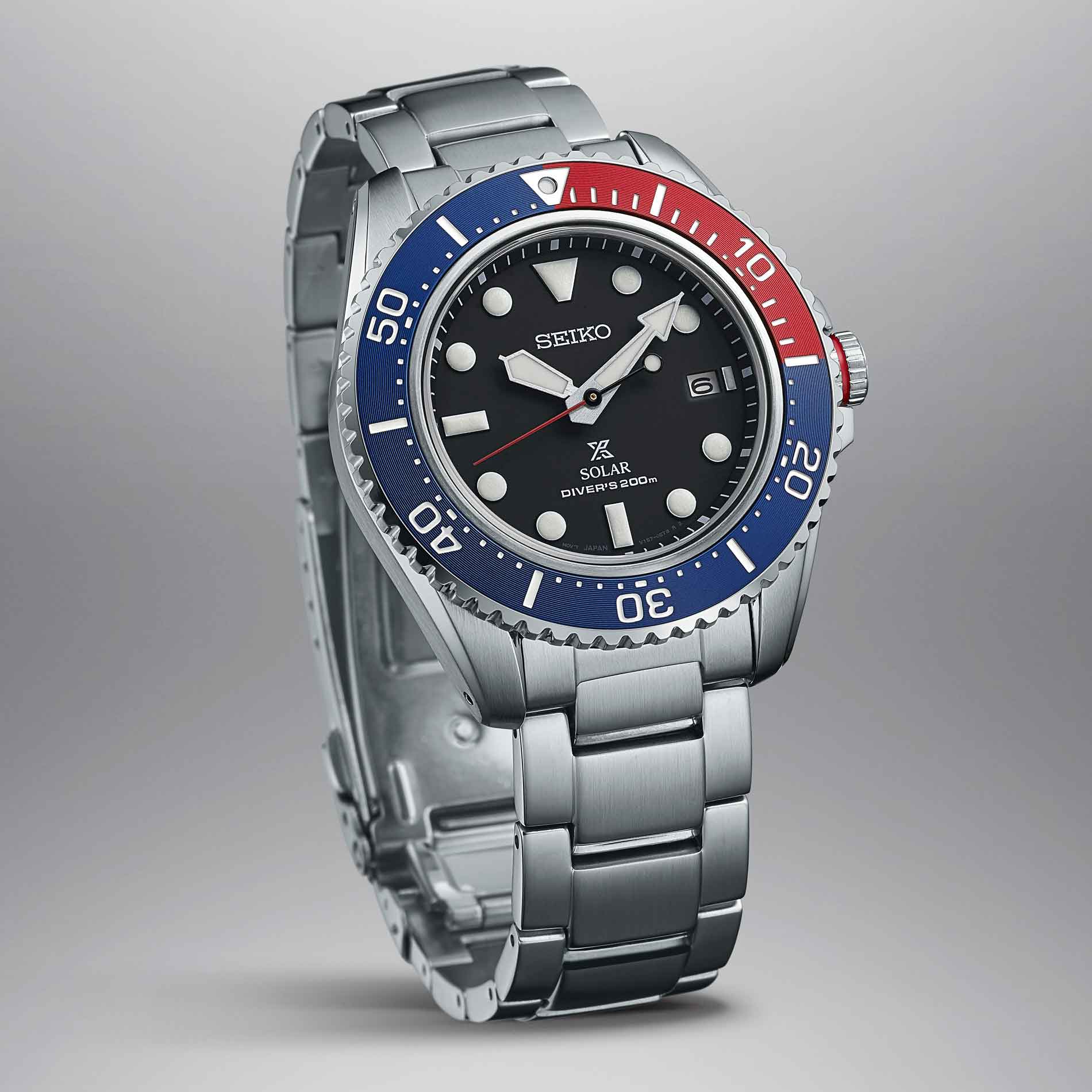 Seiko herreur i stål med sort urskive og blå / rød krans. Uret ses skråt fra siden på en grå baggrund.