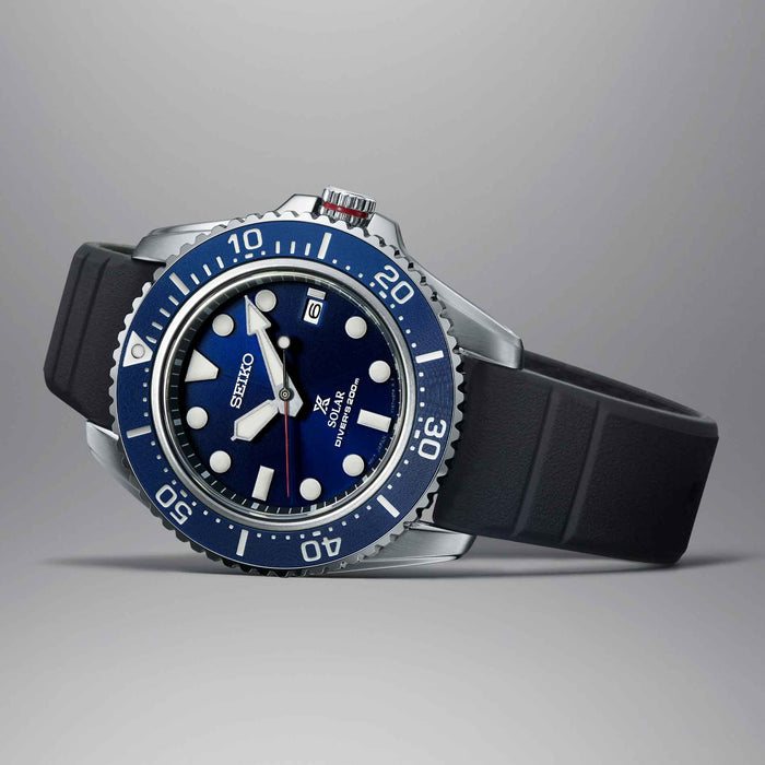 Seiko herreur i stål med blå urskive og blå krans. Uret har en sort solikonerem. Uret ses liggende på en grå baggrund.