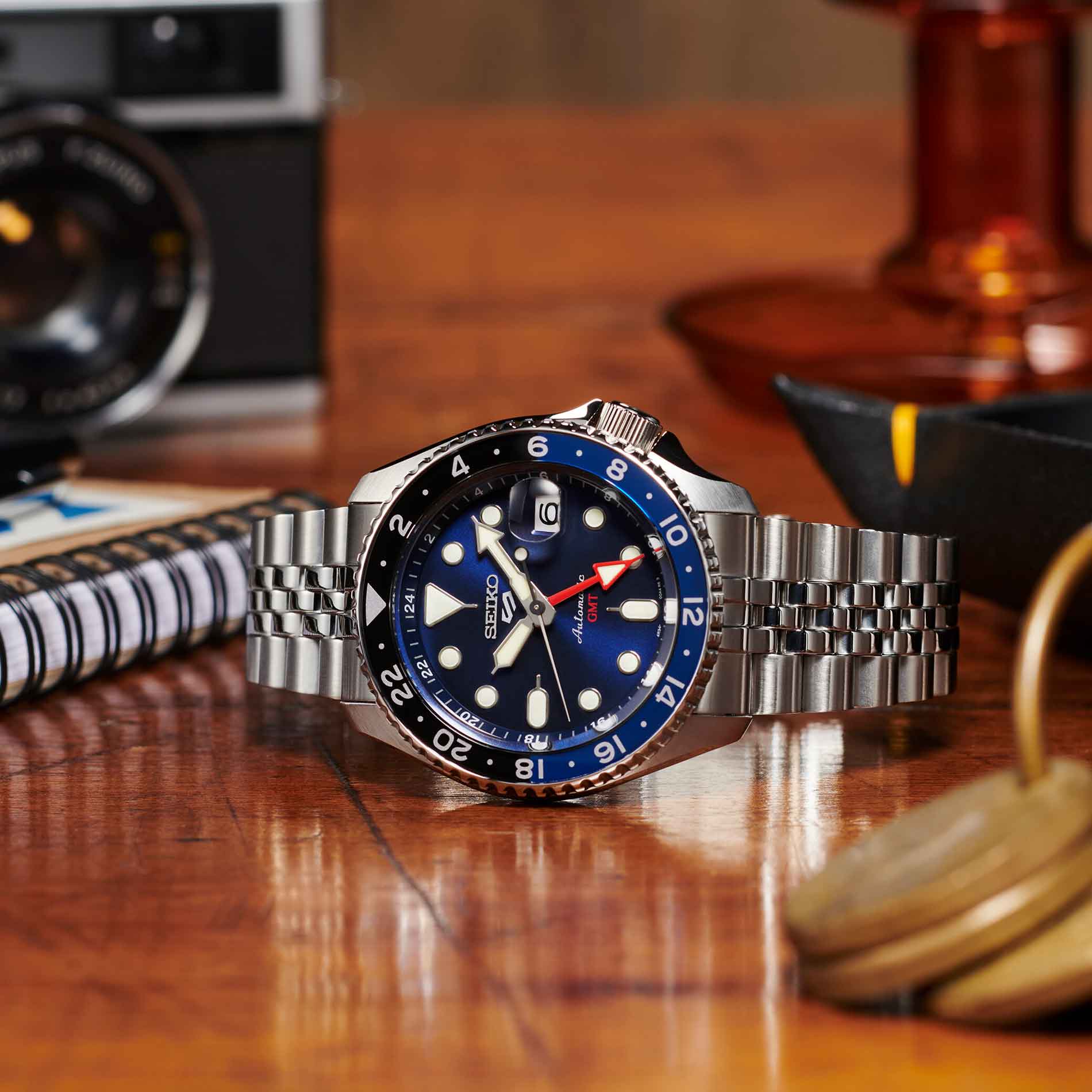 Seiko herreur i stål med blå urskive, hvide visere og indekseringer samt rød GMT-viser. Uret ses liggende på et træbord.