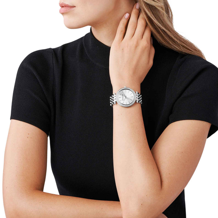 Sølvfarvet Michael Kors Darci MK3190 dameur med sølvfarvet urskive og funklende hvide sten rundt i kransen. Uret er set på håndledet af en kvinde iført en sort T-shirt.