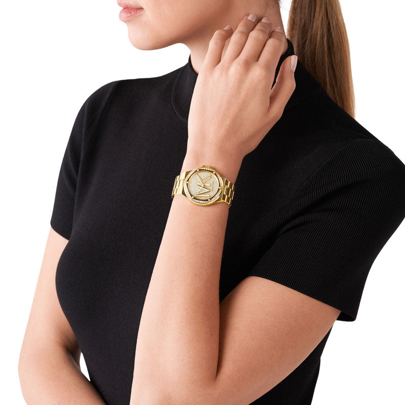 Guldfarvet Michael Kors Lennox MK7229 dameur med gylden urskive og guldfarvet lænke. Uret har mange funklende sten på skive og krans. På skiven står logoet MK. Uret ses på håndledet af kvinde iført sort T-shirt..