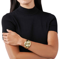 Michael Kors Slim Runway MK3178 dameur i guldfarvet rustfrit stål. Uret har en flot gylden urskive med indeksmarkeringer. Uret ses på håndledet af en kvinde iført en sort T-shirt.