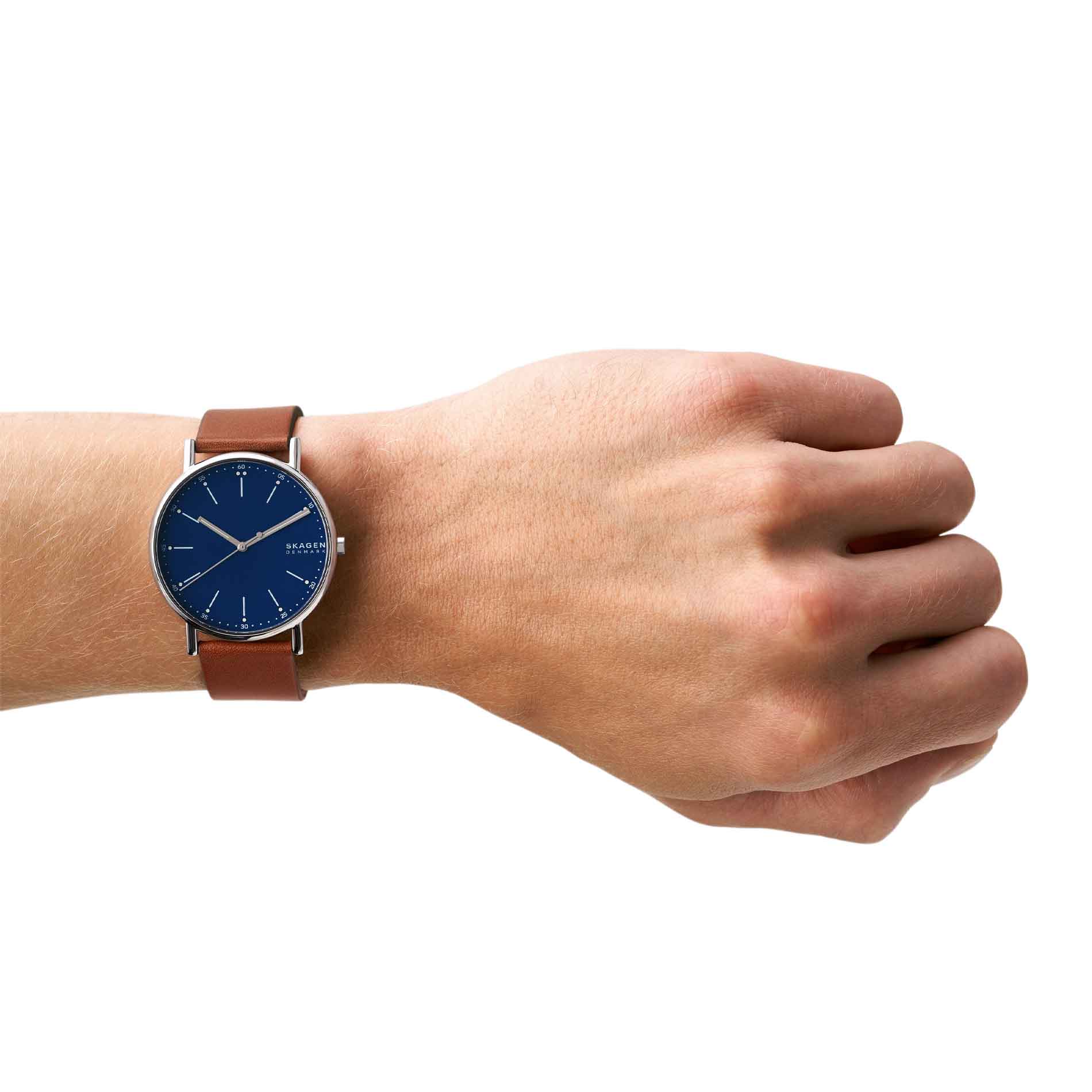 Skagen herreur med stålurkasse, blå skive og brun læderrem. Uret ses på et håndled.