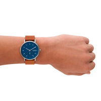 Skagen ur med stålurkasse, blå skive og brun læderrem. Uret har arabertal på skiven. Uret ses på et håndled.