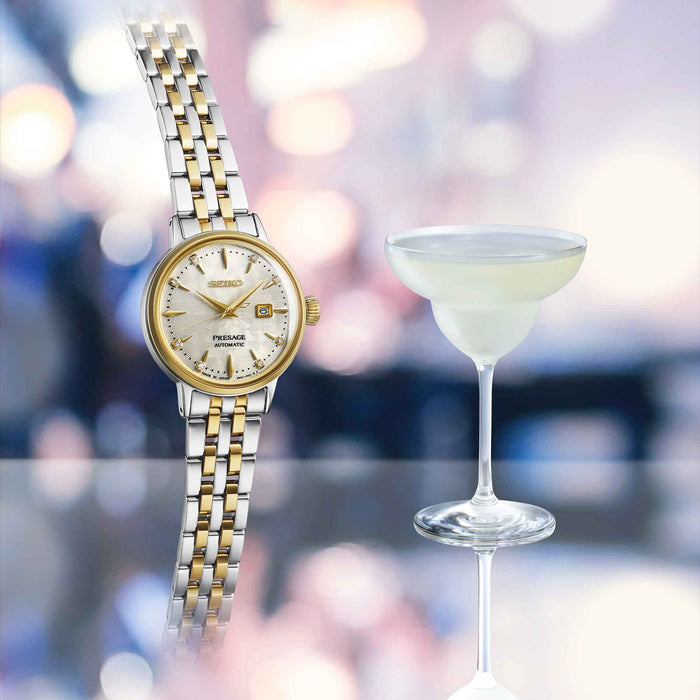 Seiko dameur i stål og guldtonet stål med champagnefarvet skive. Uret ses ved siden af en cocktail.