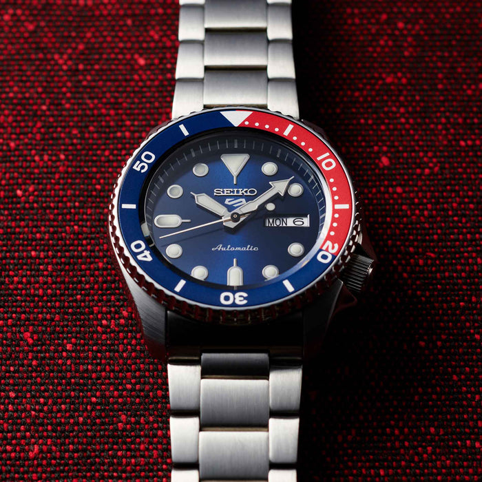 Seiko herreur i stål med blå urskive og blå / rød krans (Pepsi). Uret ses liggende på et rødt stykke tekstil.