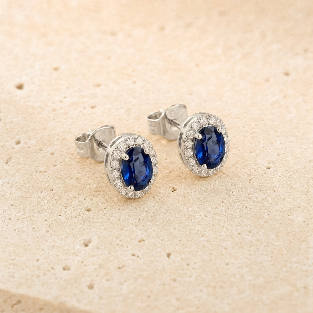 Elina Roset ørestikker i sterling sølv med dybblå oval zirkonia omgivet af klare zirkonia. Det tidløse rosetdesign er fotograferet på lys naturstensbaggrund med fokus på stenens farvedybde og sølvets elegante finish.