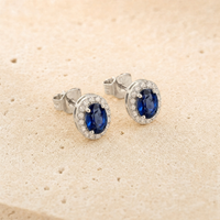 Elina Roset ørestikker i sterling sølv med dybblå oval zirkonia omgivet af klare zirkonia. Det tidløse rosetdesign er fotograferet på lys naturstensbaggrund med fokus på stenens farvedybde og sølvets elegante finish.