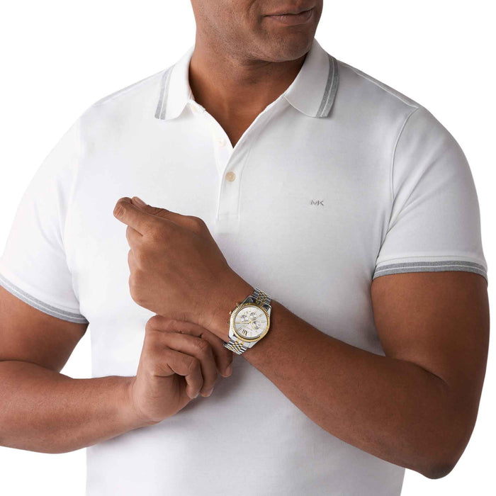 Sølvfarvet Michael Kors Lexington MK8344 herreur med guldtonede detaljer. Uret har lænke, tre trykknapper på siden, og det har romertal, indekseringer og chronographer på den grå urskive. Uret ses på håndledet af en mand iført en hvid T-shirt.