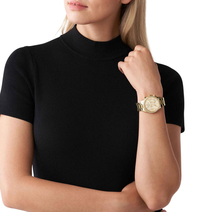 Guldfarvet Michael Kors Ritz MK6356 dameur med hvide sten i kransen. Uret har en gylden skive og en guldfarvet lænke. Uret ses på håndledet af en kvinde iført en sort T-shirt.