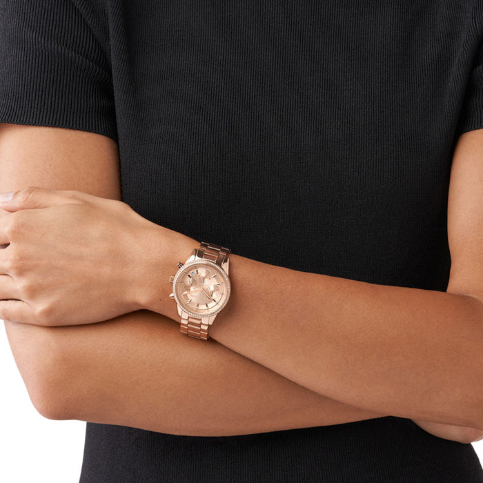 Rosafarvet Michael Kors Ritz MK6357 dameur med hvide sten i kransen. Uret har en rosa skive og en rosafarvet lænke. Uret ses på håndledet af en kvinde iført en sort T-shirt.